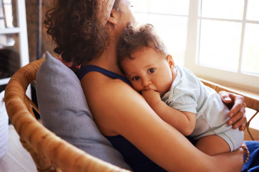Rear view of young dark skinned female sitting by window in chair, nursing nine month old toddler. Mixed race mother soothing her infant son to sleep. Childcare, family and motherhood concept